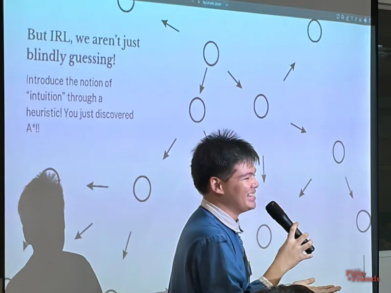 A speaker in a denim jacket laughs mid-talk, mic in hand, in front of a slide of circles and arrows.