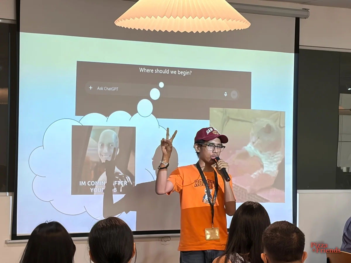 A speaker in an orange shirt and maroon cap flashes a peace sign while talking into a mic, in front of a slide that reads "IM COMING AFTER YOUR JOB", with the audience in the foreground.