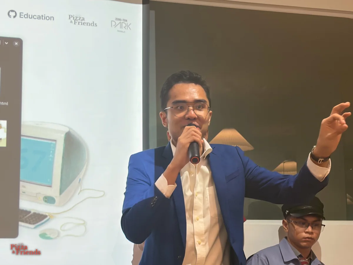 A speaker in a blue suit talks into a mic with one arm raised, while a person in a black cap sits nearby.