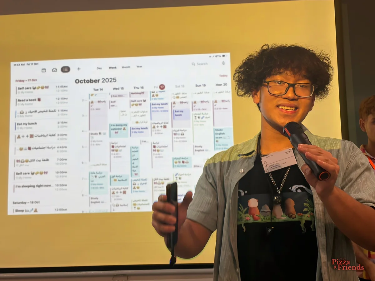 A presenter with curly hair and glasses smiles into a mic during a demo, with a weekly calendar app projected behind them.