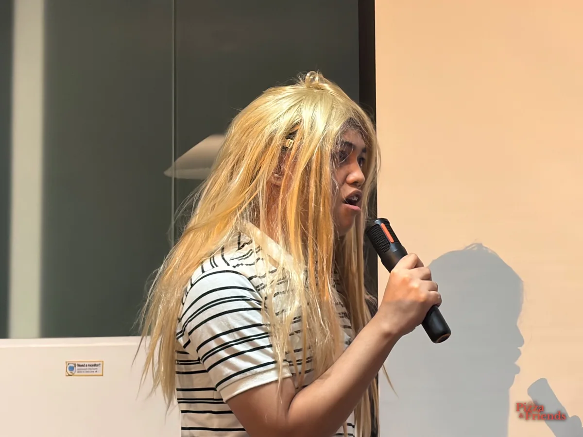 A presenter wearing a long blonde wig speaks into a mic during a pitch.